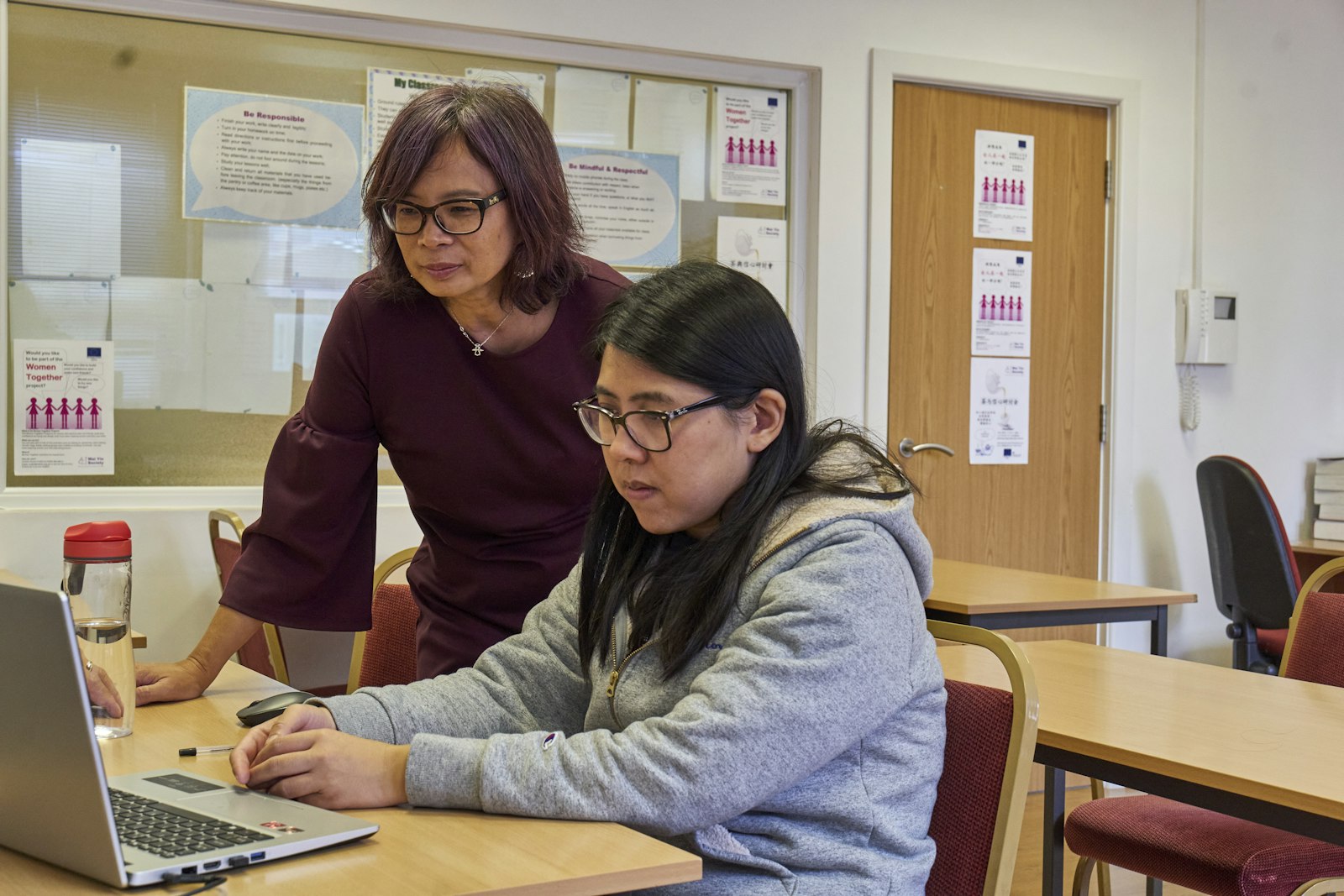 A coach standing alongside an adult participant working at a laptop, the kind of patient one-to-one support our program will provide